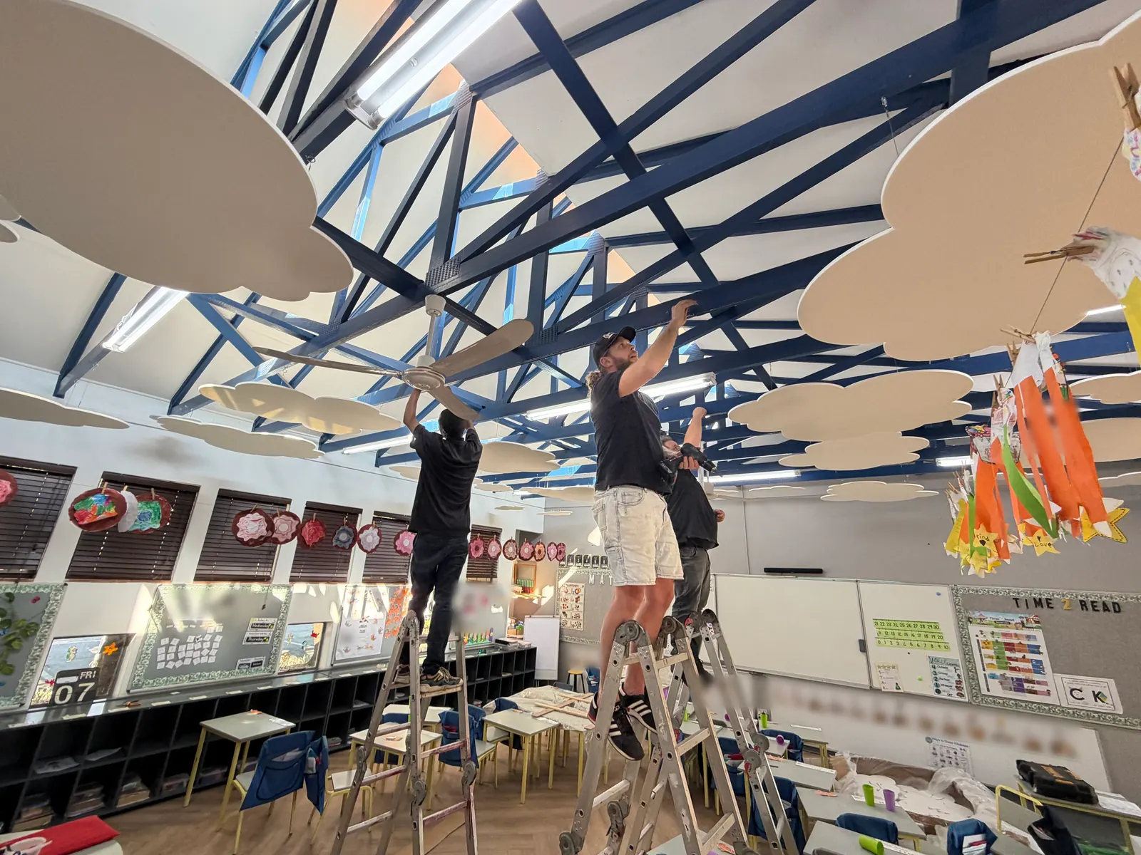 Installers on ladders hang white felt clouds under the roof trusses of a Johannesburg classroom
